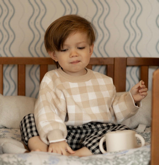 Child sitting on a bed with a checkered gingham pattern, holding a mug.