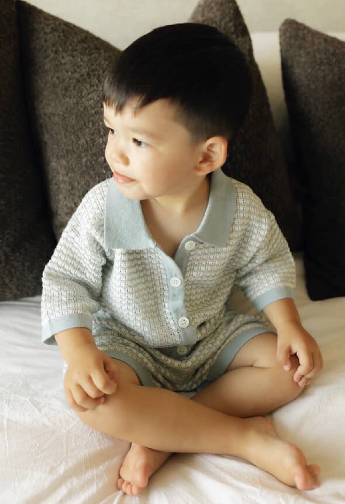 Child wearing a light blue knitted outfit sitting on a bed with dark pillows.
