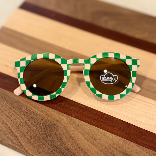 Round sunglasses with a green and cream checkered frame and dark brown lenses, resting on a striped wooden board.