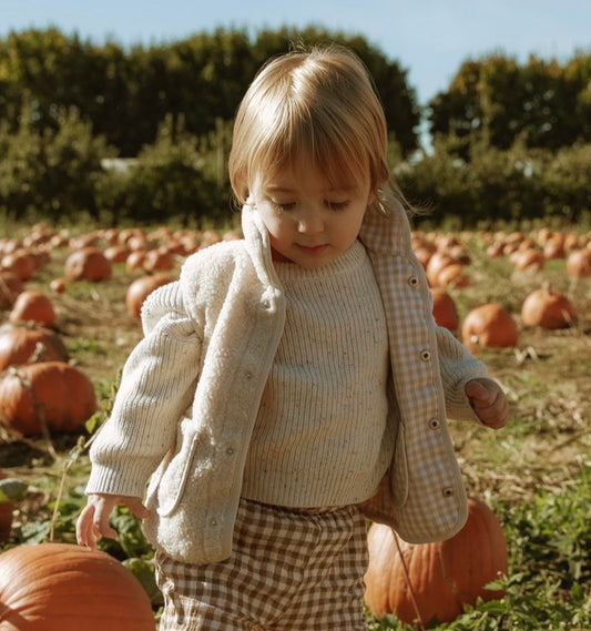 Child in a pumpkin patch wearing a checkered  gignham sherpa vest and sweater