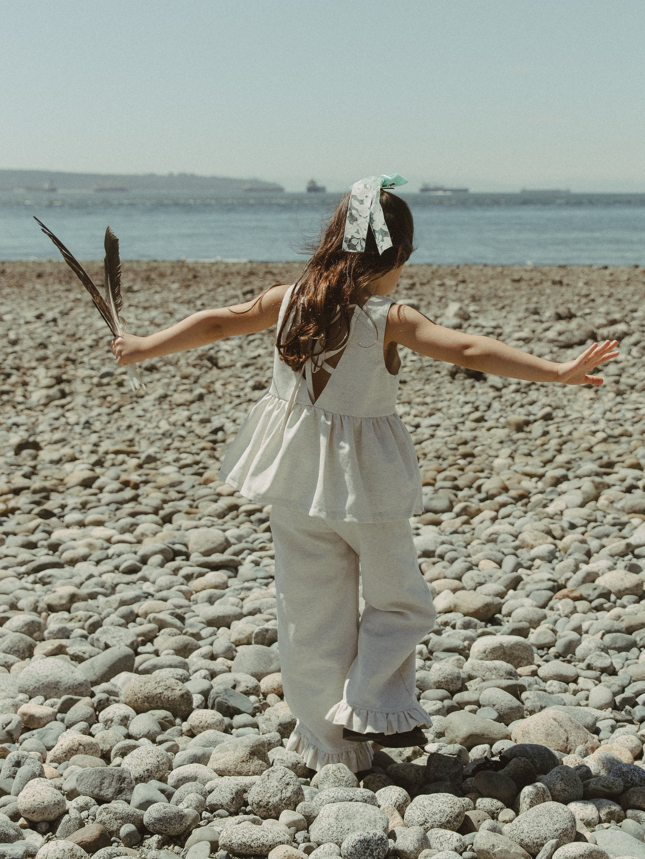 A young girl stands on a pebble beach with her arms outstretched, wearing an oat linen sleeveless ruffle top and ruffle hem oat colored pants, holding a feather in one hand. The sea and sky are visible in the background.
