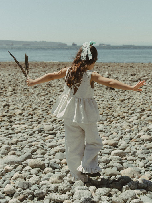 A young girl stands on a pebble beach with her arms outstretched, wearing an oat linen sleeveless ruffle top and ruffle hem oat colored pants, holding a feather in one hand. The sea and sky are visible in the background.