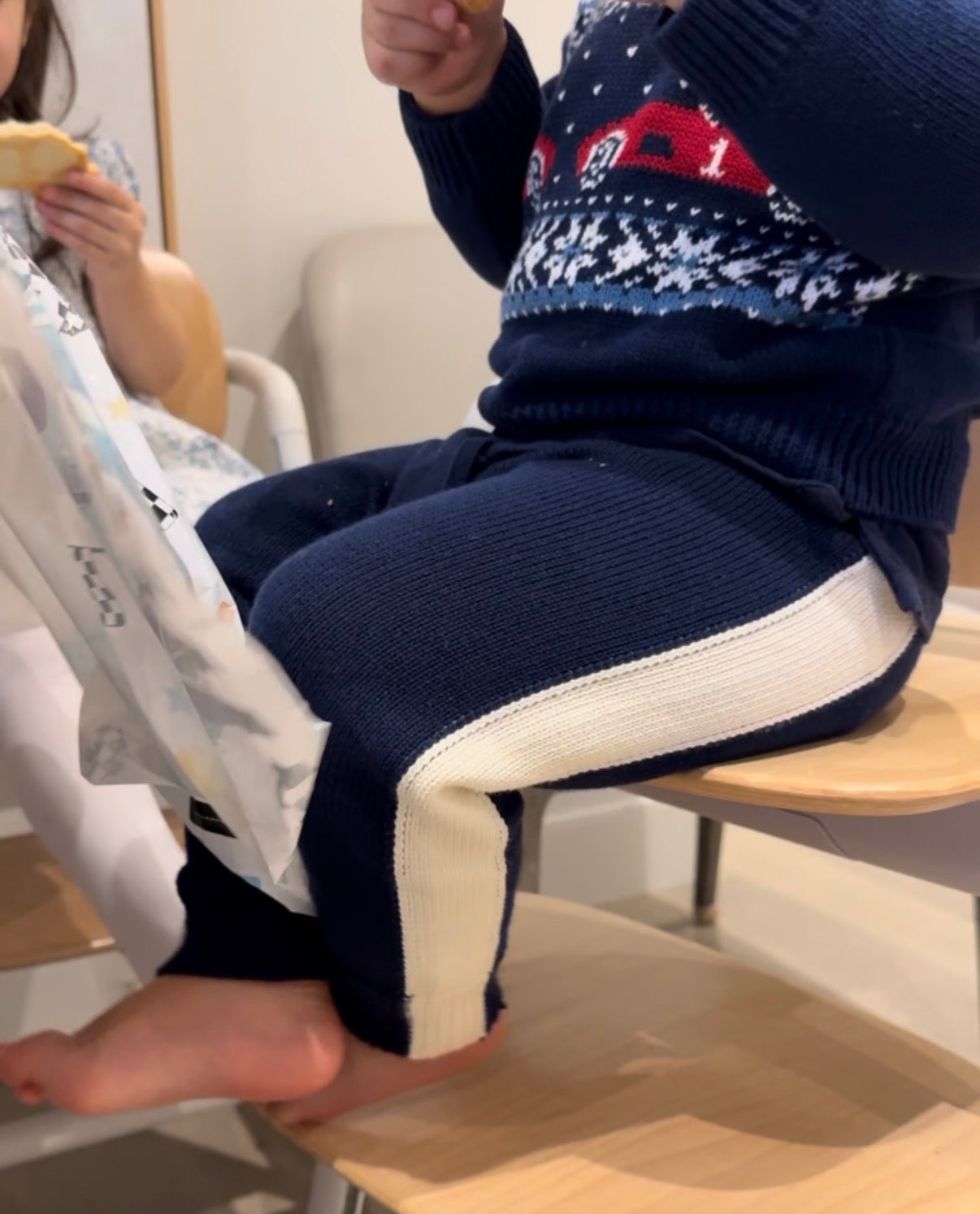 Child wearing a navy sweater with a red car design and  blue and white retro pants sitting on a chair.