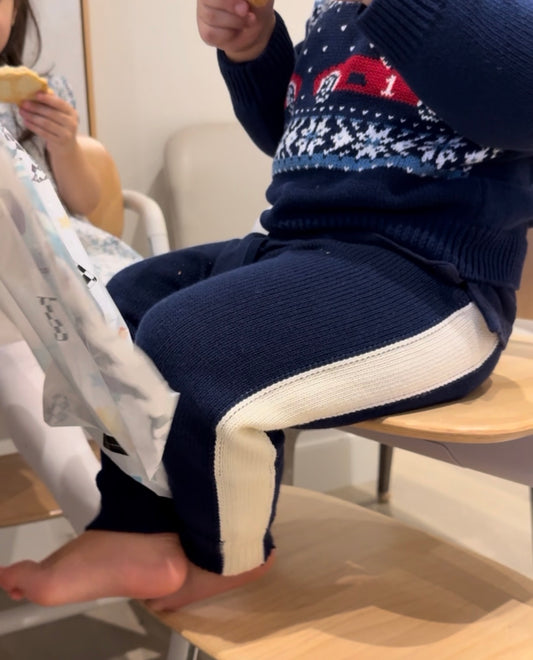 Child wearing a navy sweater with a red car design and  blue and white retro pants sitting on a chair.
