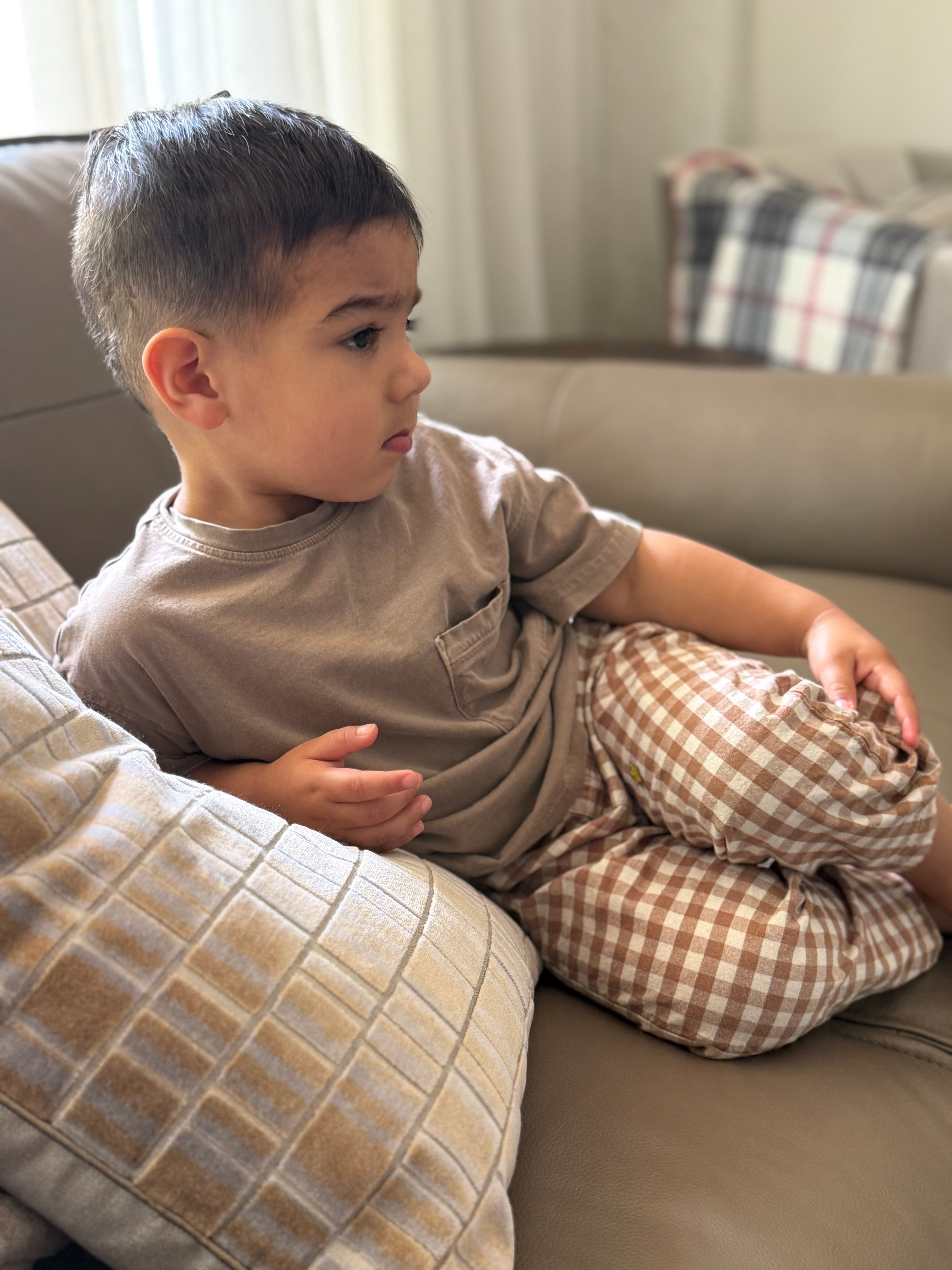 Child sitting on a couch wearing a brown shirt and checkered brown gingham pants in a living room setting.