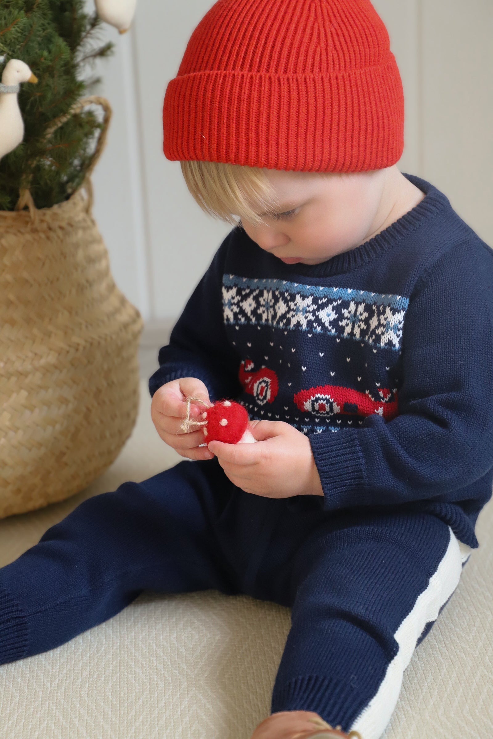 Child wearing a navy sweater with red and white patterns and a red knit hat, sitting on a light-colored surface.