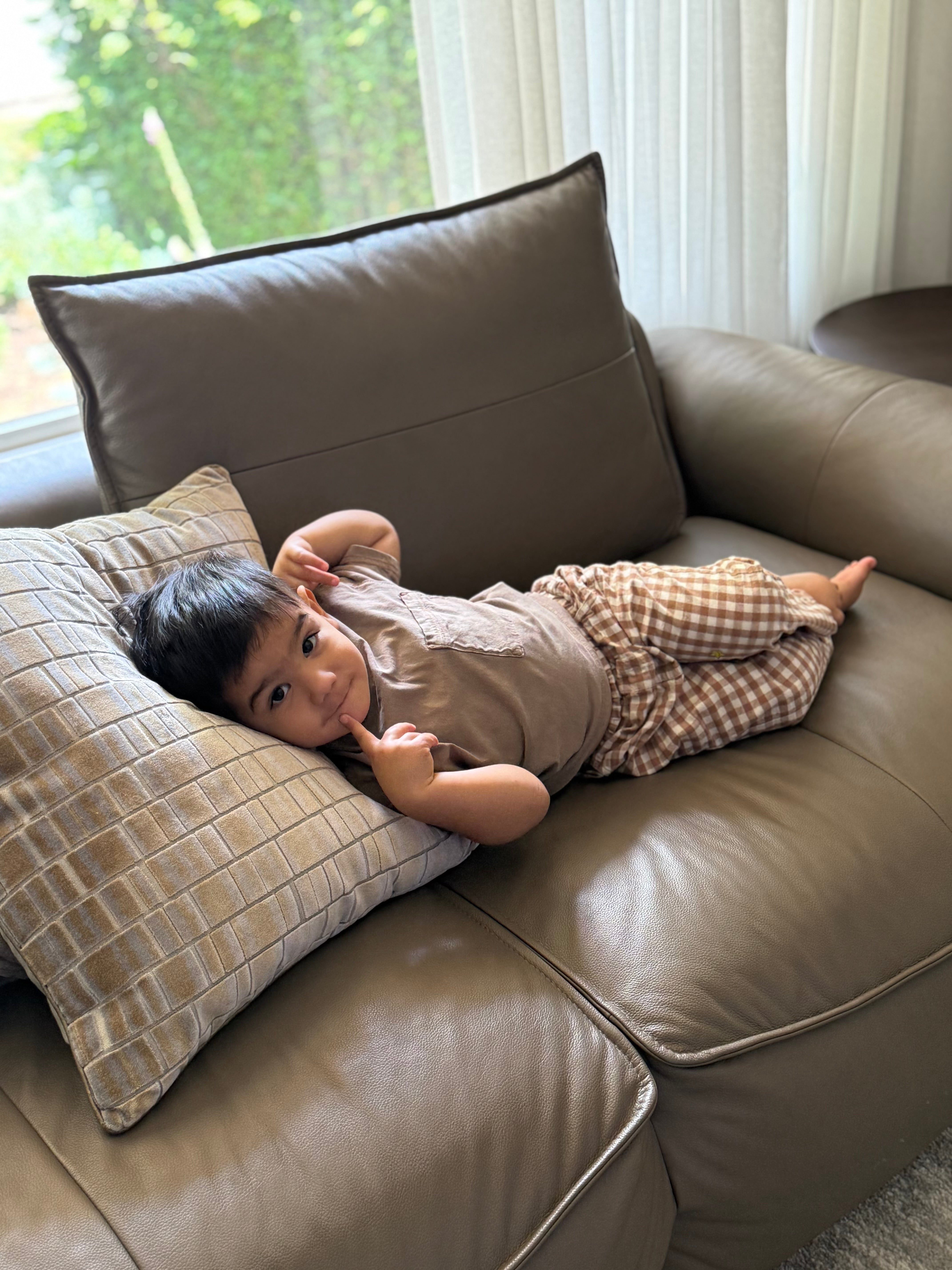 Child lying on a brown leather couch with pillows in brown gingham trouser pants 