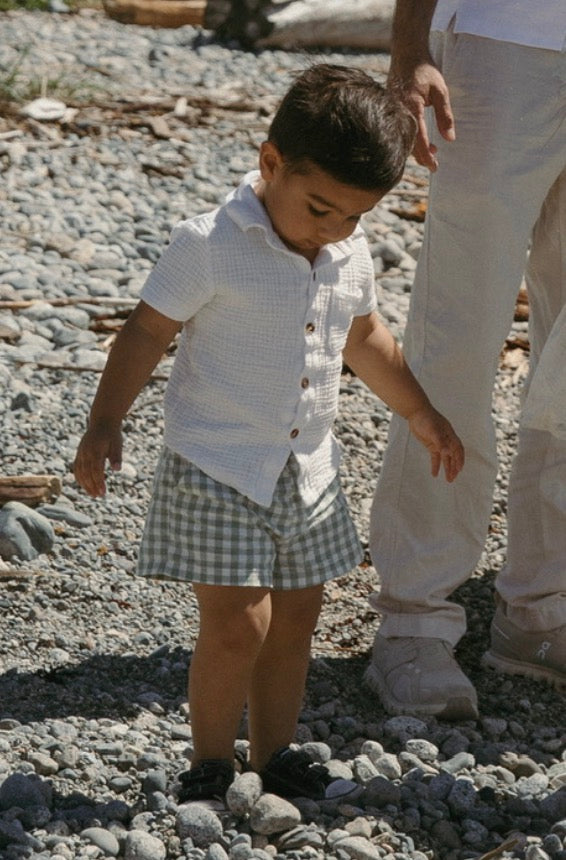 A young boy walking outdoors on a gravel surface, wearing a white short-sleeve button-up shirt and a light green and white gingham shorts. An adult in light-colored pants is partially visible beside him.