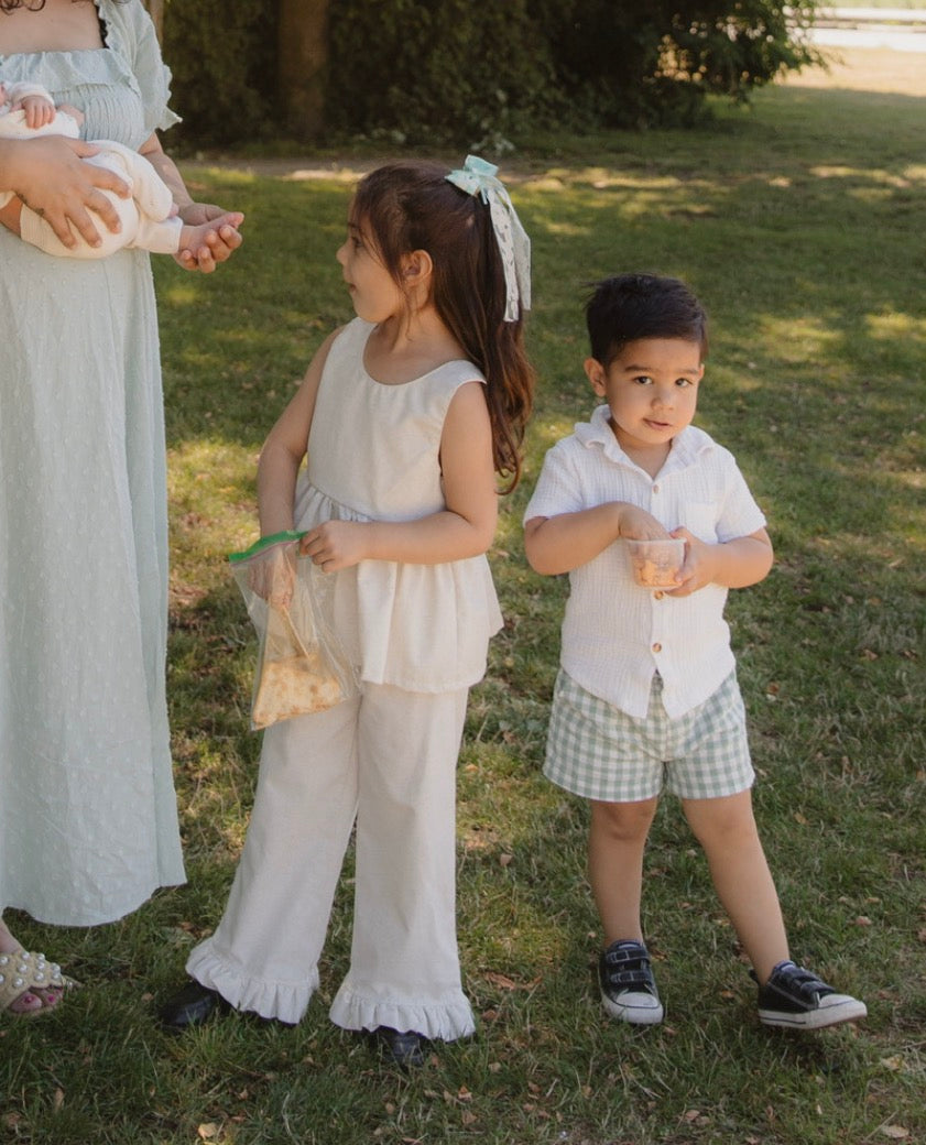 A woman and two young children standing outdoors on a grassy lawn. The children are wearing matching light-colored clothing; the girl wears a white sleeveless top and wide-leg ruffled pants, and the boy wears a white button-front shirt and green green-and-white gingham shorts.