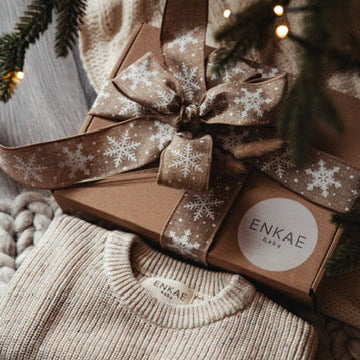 A set of Enkae Baby brand knitwear pieces, including a hat, a pair of shoes, and a pair of socks, wrapped in a box with a ribbon and snowflake pattern.