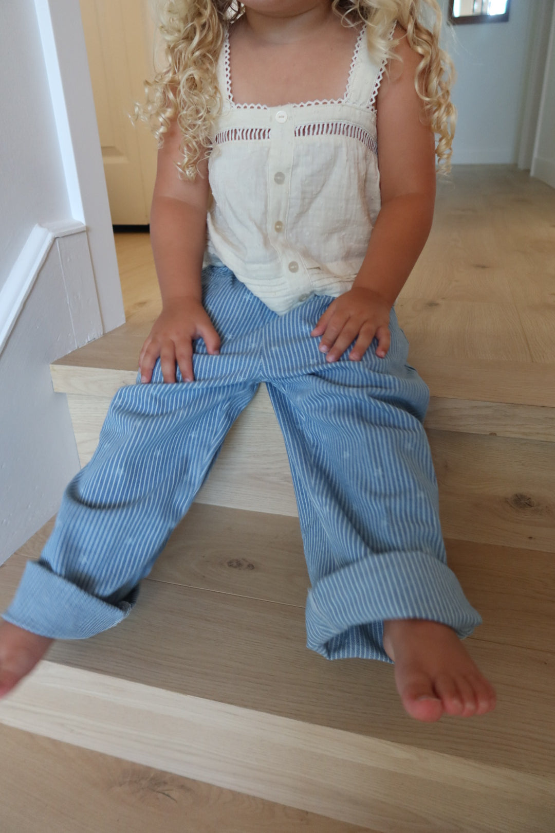 A toddler sitting on light wood stairs wearing a cream sleeveless top with crochet trim and light blue pinstriped pants.