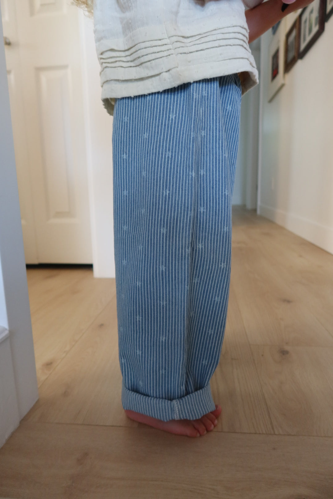 A child standing barefoot on a light wood floor in a hallway, wearing loose blue and white vertically striped pants with a small white star pattern and a cream-colored top.