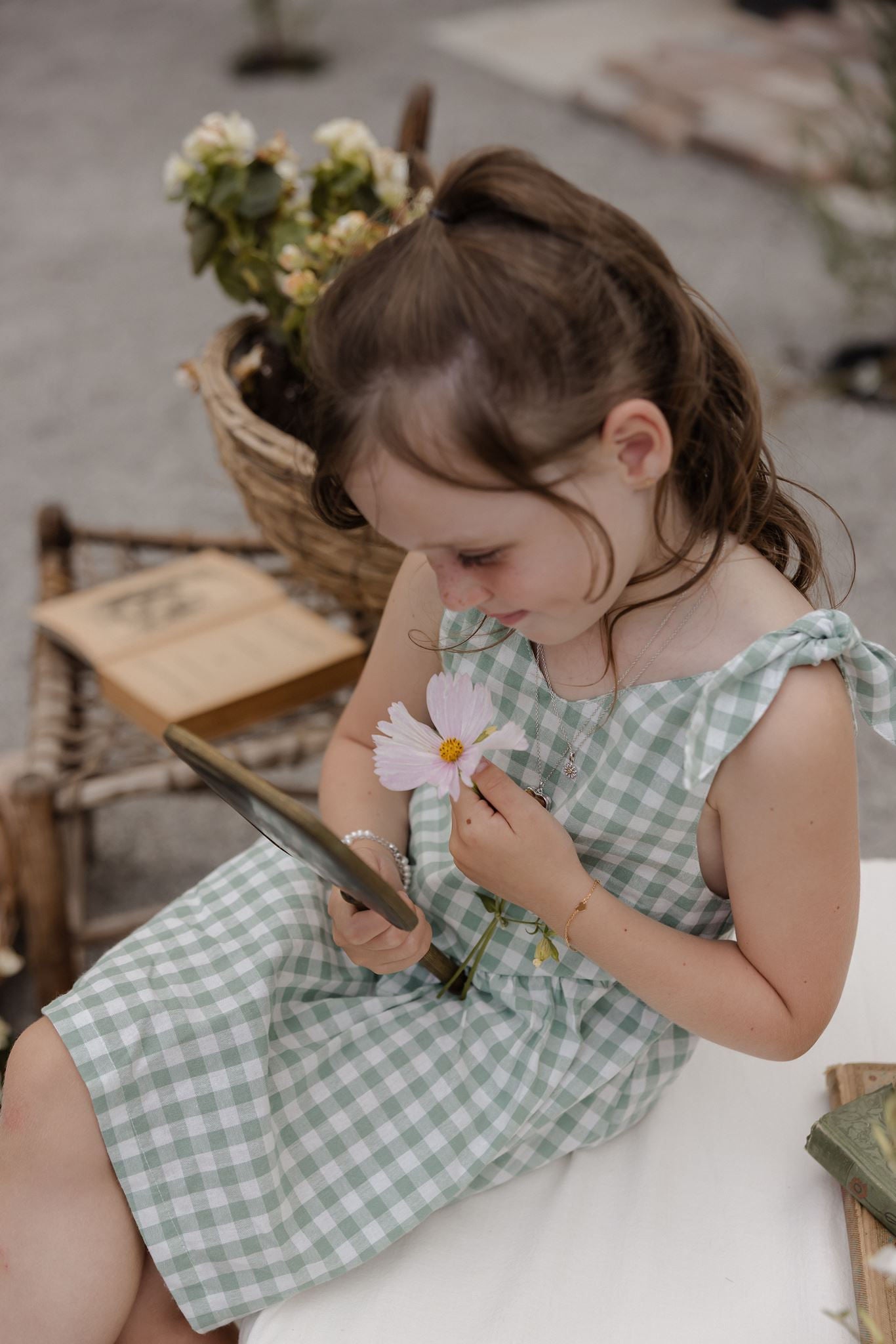 A young girl with brown hair in a ponytail sits on a white bench, wearing a light green and white gingham dress. She is holding a pink flower and looking down at it, with a rustic wicker basket and books in the background.
