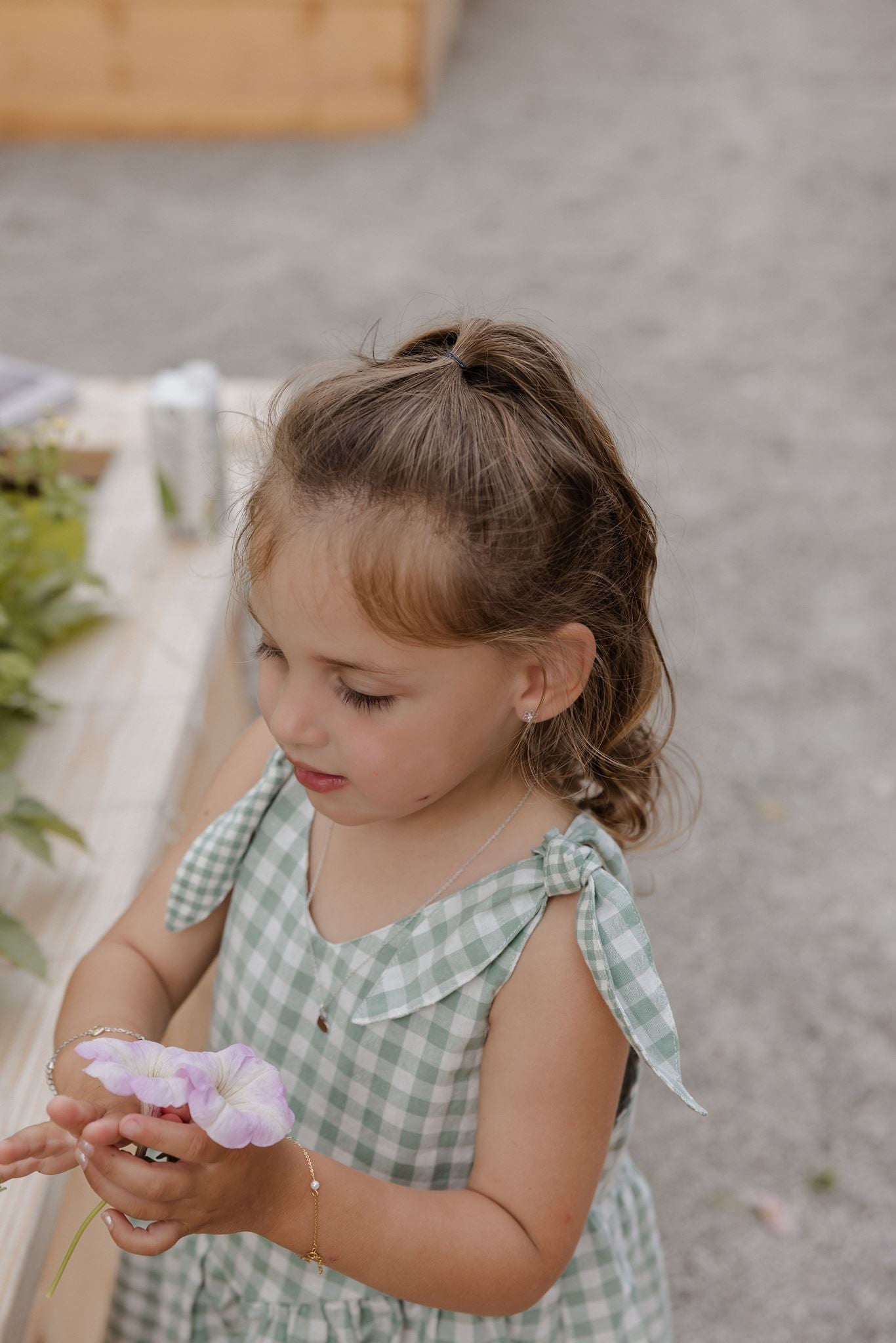 A young girl with light brown hair in a ponytail, wearing a green and white gingham dress, holding a light purple flower. She is photographed from a high angle against a soft-focus background of gray pavement and a wooden structure.