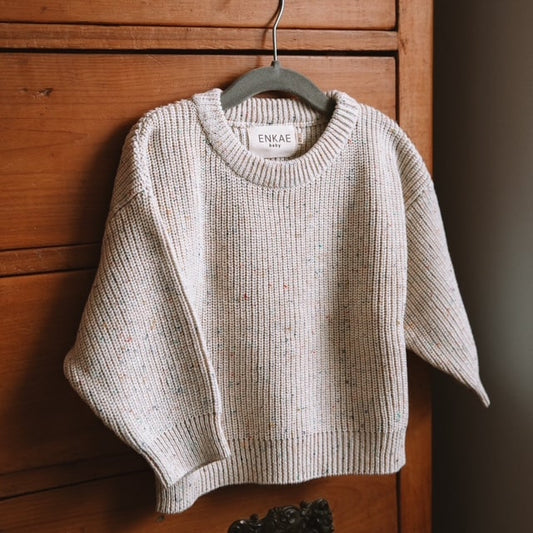 The Confetti Jumper hanging on a wooden hanger, displayed on a wooden chest of drawers. The neutral beige color with colorful specks is clearly visible, emphasizing its soft, premium cotton knit.